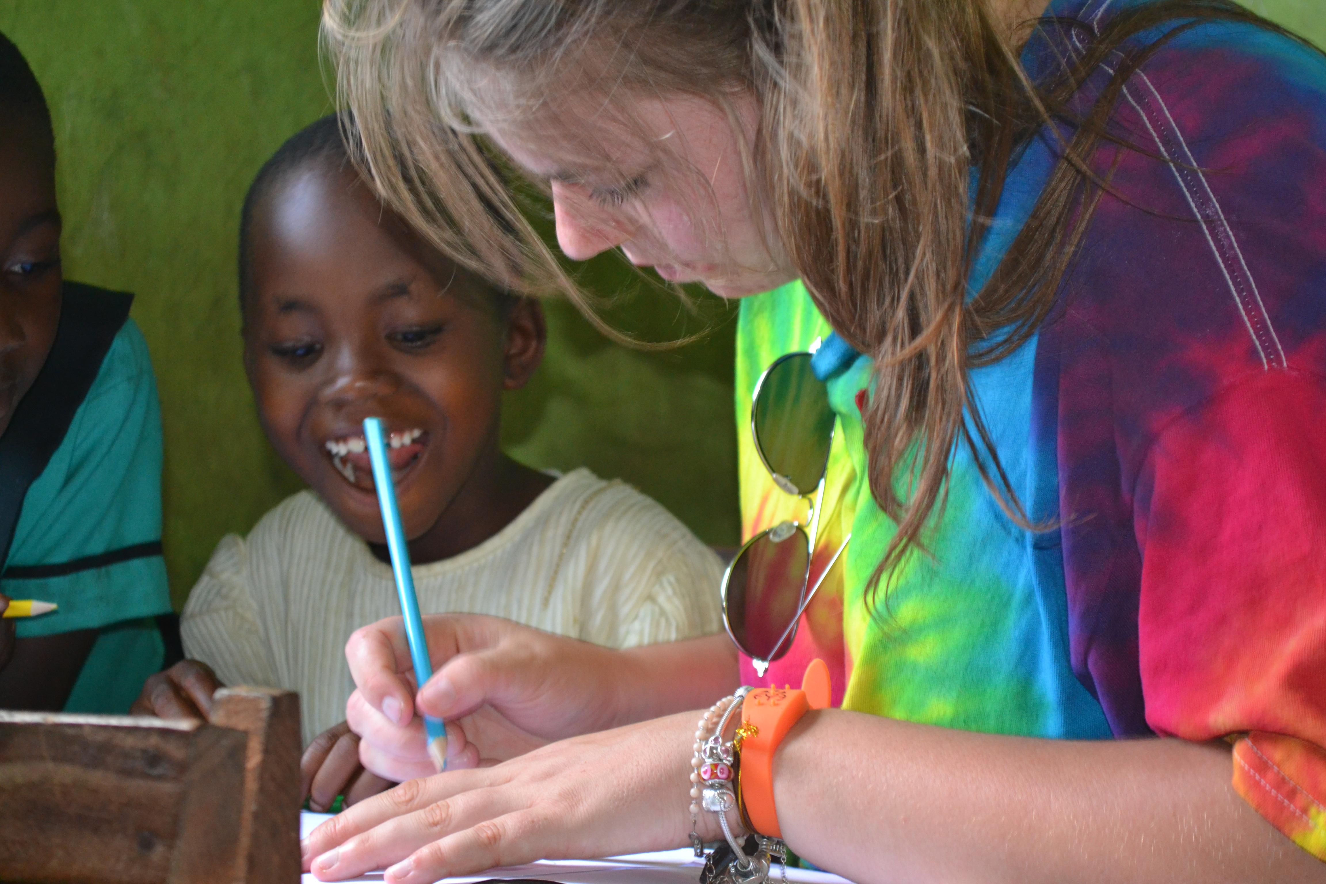 A Projects Abroad Childcare volunteer helps a young child with a writing activity at a school in Ghana. A child listens to a Childcare volunteer explain an English writing activity at a school in Ghana.